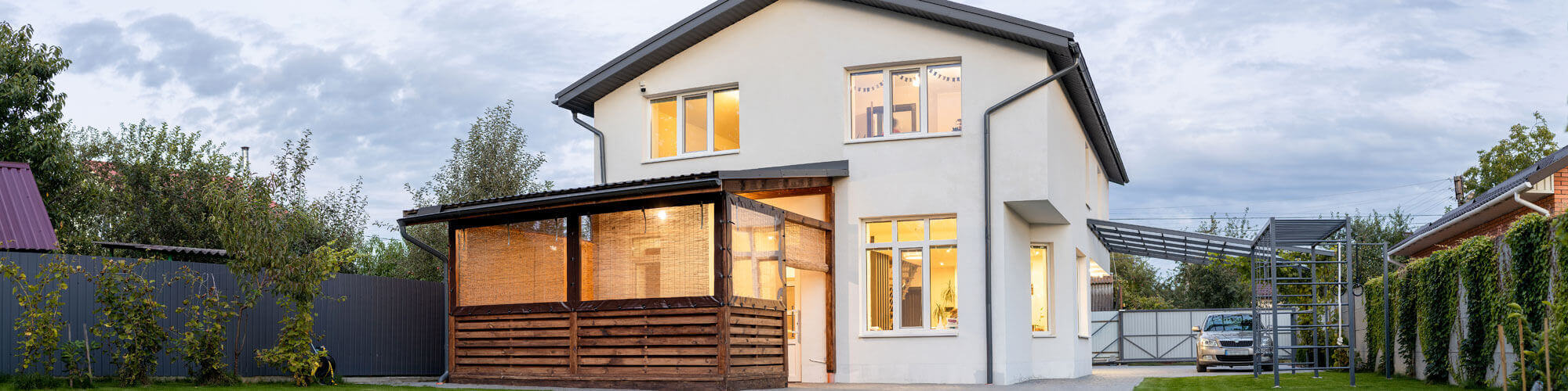Exterior View Of Modern White House With Courtyard, Patio Area, Green Grass Lawn, Garden And A Car In The Evening